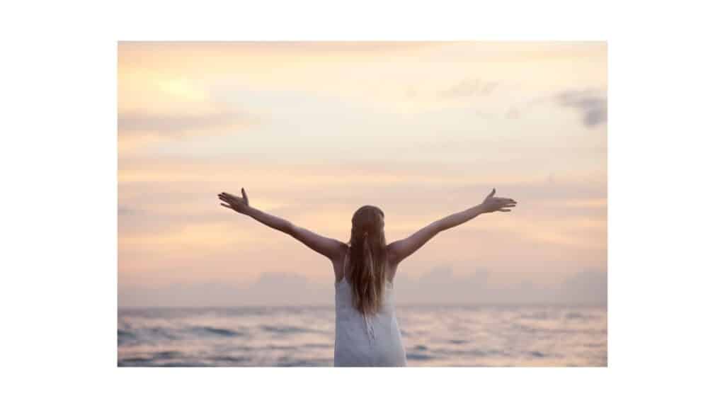 Happy Woman after ketamine therapy standing with back to reader, arms outstretched towards the ocean.